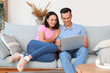 © Pixel-Shot - Happy couple using laptop while sitting on sofa in living room at home