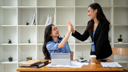 Wall Mural - Two cheerful Asian businesswomen are giving high fives to each other while working together.