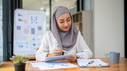 Wall Mural - An attractive Asian Muslim businesswoman wearing a hijab focuses on financial reports