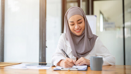 An attractive Asian Muslim businesswoman is filling out forms, signing document at her desk