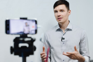 Young guy records video blog on phone camera indoors. Caucasian man conducts training remotely. Standing in front of phone. Live broadcast or streaming.