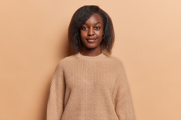 Wall Mural - Portrait of serious dark skinned woman with short hair full lips and tender expression wears casual knitted jumper focused at camera isolated over brown background. Human facial expressions.