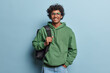 People positive emotions concept. Studio waist up of young happy smiling Hindu male student standing in centre isolated on blue background wearing green hoodie and jeans with black bag on shoulder