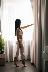 Wall Mural - Single woman is a beautiful Caucasian woman in pajamas without standing at the window of a house or in a hotel room and looking at the street on a sunny day