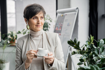 Poster - mature businesswoman with coffee cup listening music in earphone and looking away in office