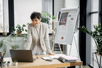 Poster - smiling middle aged businesswoman near laptop and flip chart with graphs, video call, conference