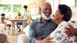 © Monkey Business - Senior Couple At Home Looking At Laptop Together With Multi-Generation Family In Background