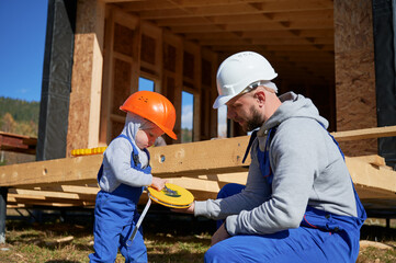 Wall Mural - Father with toddler son building wooden frame house in Scandinavian style barnhouse. Boy helping his daddy, playing with tape measure on construction site on sunny day. Carpentry and family concept.