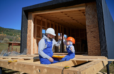 Wall Mural - Father with toddler son building wooden frame house. Male builder giving high five to kid on construction site, wearing helmet and blue overalls on sunny day. Carpentry and family concept.