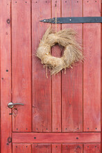 Old, Wooden, Red Door With A Metal Handle. A Round Wreath Of Dry Grass Tied With Hemp Rope Hangs On A Rusty Nail.