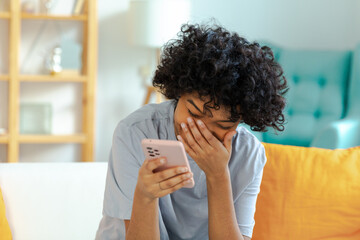 Wall Mural - Excited happy young black african american woman holding cell phone laughing feeling joy getting mobile message. Overjoyed girl laughing aloud sitting on couch watching funny video reading news