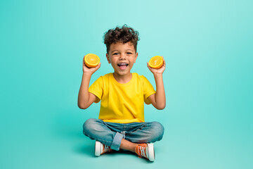 Full length photo of good mood small boy with brown hair wear stylish t-shirt sit hold sliced orange isolated on teal color background