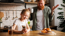Smiling Freckled Child Using Smartphone Near Blurred Angry Father During Breakfast In Kitchen, Parenting Struggles Concept