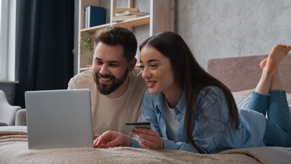 Caucasian couple family lying together on bed use credit bank card enter code in laptop computer banking app online payment smiling woman and man shopping internet order pay delivery at home bedroom