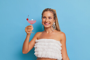 Wall Mural - Happy young woman with dreads dressed in white top having good time on blue background with glass of cocktail in hand, happy time concept, copy space