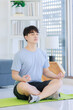 © Bangkok Click Studio - Asian young handsome fit male athlete teenager in sportswear sneakers sitting closed eyes meditating on yoga mat with lotus posture exercising training together in living room at home.