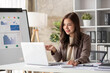 © wichayada - Cheerful business lady working on laptop in office, Asian happy beautiful businesswoman in formal suit work in workplace. Attractive female employee office worker smile.
