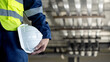 © Summer Paradive - Safety workwear concept. Male hand holding white safety helmet or hard hat. Construction worker man in protective suit and reflective green vest standing with building piping system in the background