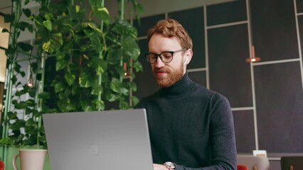 A young red-haired guy, a programmer or entrepreneur in glasses, works in a stylish cafe behind a laptop. Freelancer works remotely. Online communication. Small business.