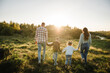 Mom, dad, daughter and son walk back in green grass in field. Happy young family with children spending time together, running outside, go in nature, on vacation, outdoors. Concept of family holiday.