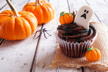 Poster - Spooky Halloween chocolate cupcake with ghost and pumpkins. Close up table scene against a white wood background.