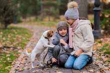 Caucasian Boy And Girl Posing Sitting On Sidewalk With Jack Russell Terrier Dog In Park In Autumn.