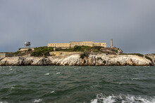 San Francisco, CA, USA - July 12, 2023: Alcatraz Island West Rocky Shite-guano Covered Cliffs. Beige Prison Building And Water Tower On The Left. Gray Foggy Sky And Greenish Bay Water
