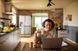 © Marko Geber - Middle aged mixed ethnicity woman drinking tea while working and using her laptop in the kitchen at home
