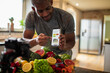 © Marko Geber - Middle aged african american male food nutritionist and blogger promoting and showing his dieting app to his customers and clients while recording in the kitchen with organic fruits and vegetables
