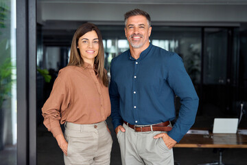 Smiling Latin middle aged business man and woman in office, portrait. Two happy confident professional mature corporate executive leaders company managers standing in office looking at camera.