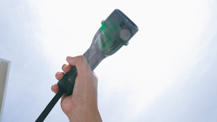 Closeup hand grasping an EV plug for electric vehicle with the midday sky in the background as progressive idea of alternative sustainable clean and green energy for environmental concern.