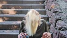 An Informal Cheerful Teen Girl Actively Shakes Her Head While Sitting On The Steps Outside. Young Blonde Girl Waving Her Hair.