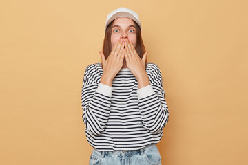 Wall Mural - Surprised astonished woman wearing baseball cap and striped shirt isolated over beige background looking with big eyes covering mouth with palms