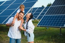 Young Family With A Small Child In Her Arms On A Background Of Solar Panels. A Man And A Woman Look At Each Other With Love. Solar Energy Concept.