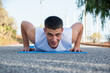 Young Caucasian man does push-ups on his athletic mat in a public park. He smiles at the camera.