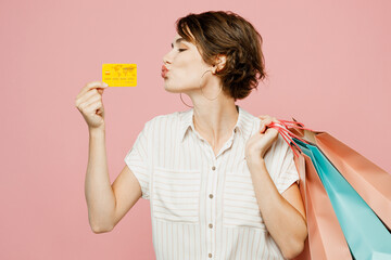 Wall Mural - Young fun woman wear casual clothes hold shopping paper package bags, kiss mock up of credit bank card isolated on plain light pastel pink color background studio. Black Friday sale buy day concept.