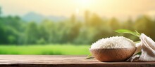 Jasmine Rice In Wooden Bowl On Vintage Wooden Desk Table With Green Rice Field And Sunlight In Background