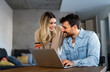© NDABCREATIVITY - Cheerful young couple using laptop and smiling while shopping online at home