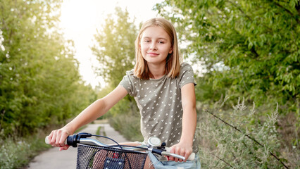 Wall Mural - Cute preteen girl with bycicle outdoors looking at camera and smiling. Pretty child with bike at city street at summer park