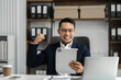 © NINENII - Happy young businessman looking at successful excited tablet sitting at desk raising hands in yes gesture celebrating business success.