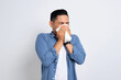 Unhealthy young Asian man in casual shirt blowing nose in paper tissue isolated on white background. Influenza virus concept