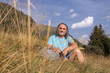 © Guzel - Mature man sitting in tall grass with backpack and smiling at the camera against the backdrop of mountains and sky