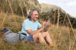 © Guzel - A gray-haired long-haired man with a backpack and smartphone sits in grass against the backdrop of mountains