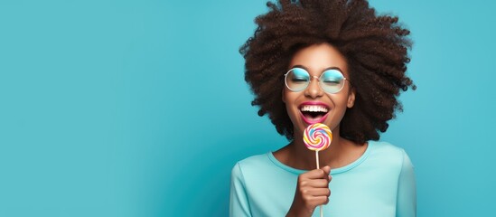 Happy African American woman in casual clothes holding a candy lollipop Black lady with a cheerful expression hiding her eye with a lollipop in a blue studio with room for text Enjoying sweets