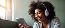 Happy Black woman using smartphone and headphones playing games at home excited African American female enjoying virtual entertainment sitting on floor in living room