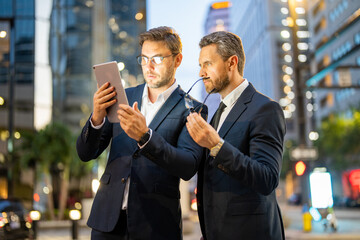 Business man standing in front of night city. Business man wearing modern suit in building night city background. The two business teams men team using tablet laptop in night city.