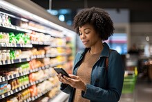 A Middle Aged Woman Is Shopping At A Grocery Store. A Middle Aged African American Woman With A Smart Phone Checks The List Of Necessary Products In A Grocery Or Supermarket With A Sale And Discounts
