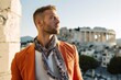 Photography in the style of pensive portraiture of a grinning boy in his 30s wearing a bold statement necklace in front of the acropolis in athens greece. With generative AI technology