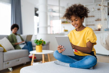 Carefree smiling African American young girl with curly hair using digital tablet sitting with crossed legs at sofa in living room with her mother in background who is using laptop.