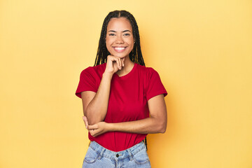 Wall Mural - Young Indonesian woman on yellow studio backdrop smiling happy and confident, touching chin with hand.